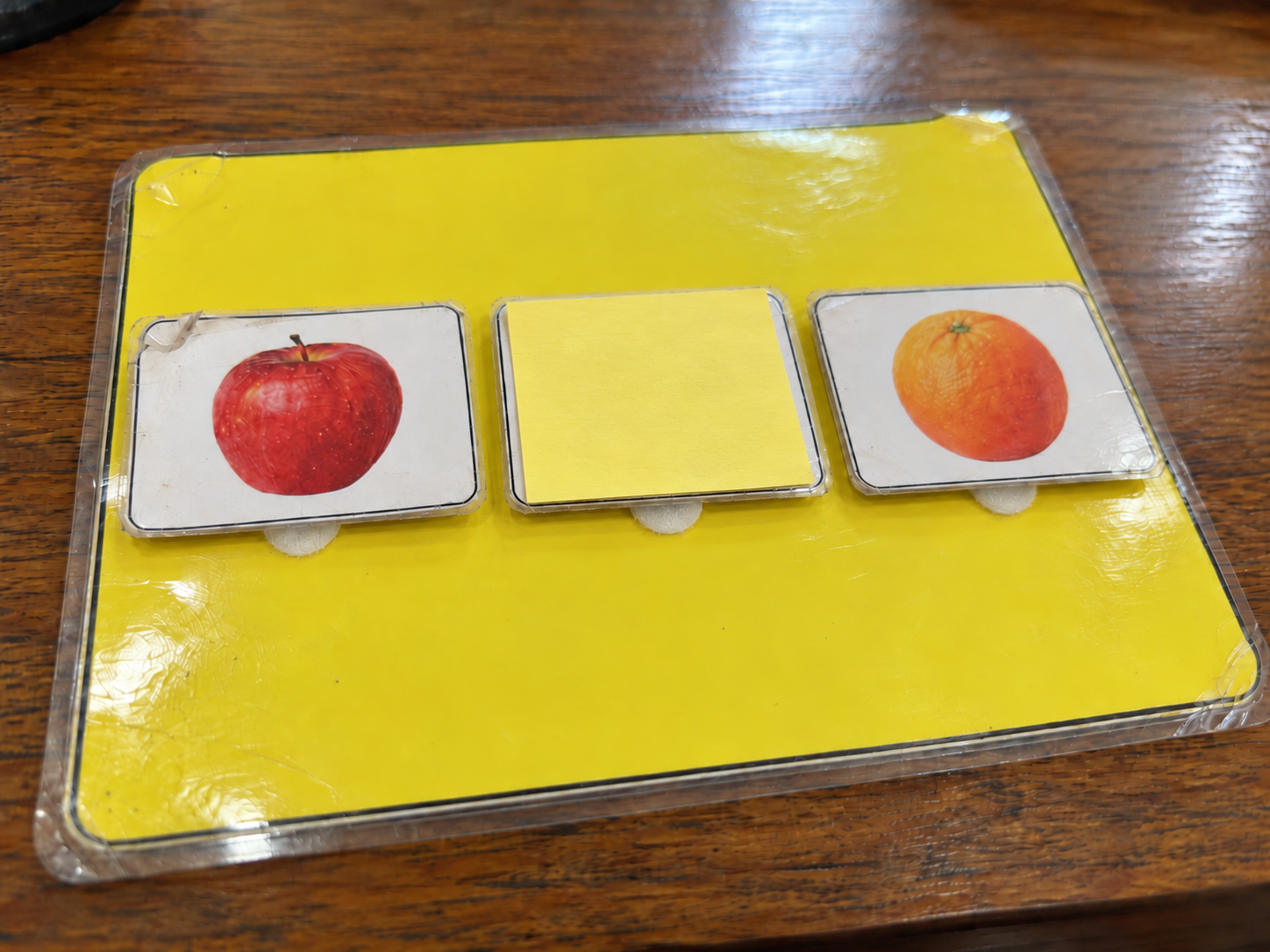 Laminated stimulus board with a yellow sticky note covering the center picture card between apple and orange cards.
